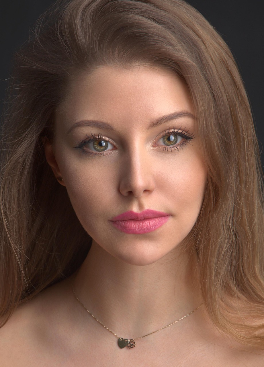 Portret młodej kobiety o długich brązowych włosach i zielono-brązowych oczach, ubranej w delikatny naszyjnik z ozdobnymi zawieszkami.